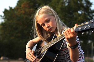 Ein junges Mädchen mit blondem Haar spielt im Freien eine richtige Gitarre, wobei sie sich auf ihre Finger konzentriert, während sie die Saiten anschlägt und drückt. Die Bäume im Hintergrund sind unscharf.